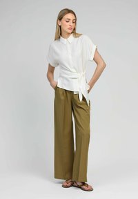 Femme portant une blouse blanche à manches courtes avec des boutons et un nœud sur le côté, associée à un pantalon large olive et des sandales marron, les mains dans les poches.