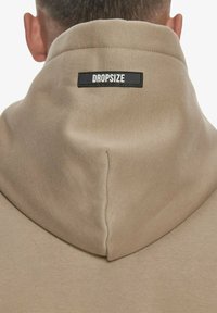 Hanorac bej cu glugă, având un etichete neagră proeminentă cu "DROPSIZE" pe spate. Material neted, texturat, cu un design structurat.