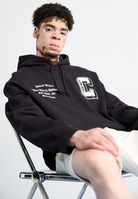 Felpa nera con cappuccio, decorata con testo bianco e un grande logo "C"; indossata sopra pantaloni corti chiari; seduto su una sedia in cromo contro uno sfondo neutro.