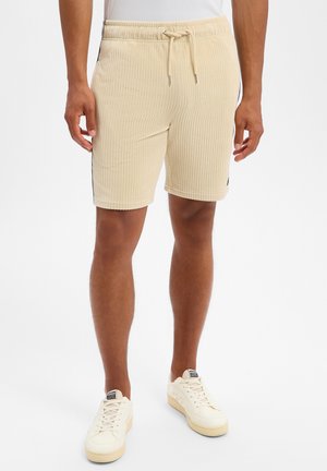 Man draagt beige geribbelde shorts met trekkoord en witte sneakers en staat tegen een effen achtergrond.