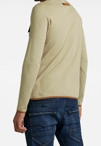 Helles Beige Langarmshirt aus strukturiertem Stoff mit brauner Saum. Die Rückseite hat ein hellbraunes Etikett mit Logo. Getragen mit blauen Denim-Jeans.