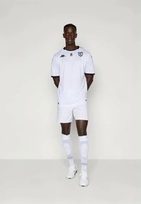 Hvitt fotballsett med kortermede skjorter og shorts, med svarte detaljer. Inkluderer knelange sokker og sportsko. Enkelt design med en logo.