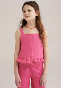 Rozā divdaļīgs tērps, kas sastāv no struktūrētas bezsleeve blūzes ar pogām un čokuroties apakšējo malu, kas papildināta ar saskaņotām biksēm.