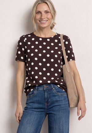 Mujer sonriente con blusa marrón de manga corta con lunares, vaqueros azules y bolso tote beige, de pie contra una pared clara.