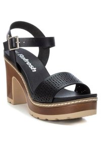 Sandalias de tacón alto de cuero negro con parte superior perforada, correa ajustable para el tobillo y suela de plataforma marrón gruesa con costuras en crema.