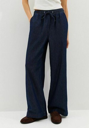 Vrouw draagt donkerblauwe wijde denimbroek met elastische tailleband en trekkoord, bruine loafers en een witte top tegen een witte achtergrond.