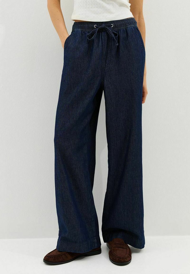 Woman wearing dark blue wide-leg denim pants with elastic waistband and drawstring, brown loafers, and white top against white background.