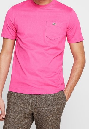 Mężczyzna noszący jasnoróżowy t-shirt z krótkim rękawem, kieszenią na piersi i małym logo krokodyla, w połączeniu z brązowymi spodniami w wzory.