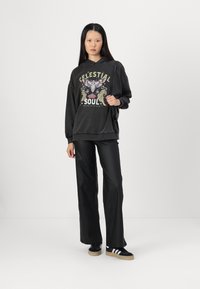 Schwarzer oversized Hoodie mit Grafikdruck, kombiniert mit schwarzen Fake-Lederhosen und schwarzen Sneakers mit weißen Streifen.