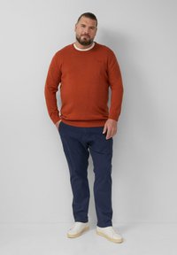 Man met een baard, gekleed in een roestkleurige trui, marineblauwe broek en witte sneakers, staande tegen een lichtgrijze achtergrond.