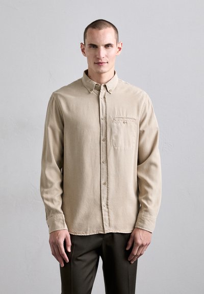 Chemise à boutons beige clair en tissu doux, dotée d'une poche poitrine, de manches longues et d'un col, associée à un pantalon foncé.