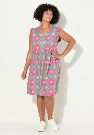 Robe rose avec des motifs géométriques verts, décolleté en V, manches courtes à volants et une silhouette fluide, portée avec des chaussures blanches.