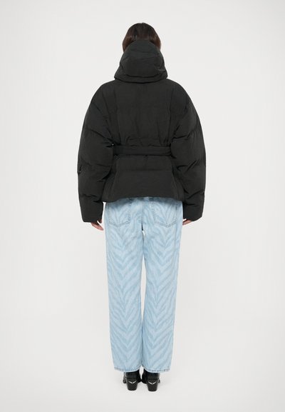 Personen står med ryggen til iført en sort dunjakke med hætte, lyseblå mønstrede jeans og sorte hæle mod en ensfarvet hvid baggrund.