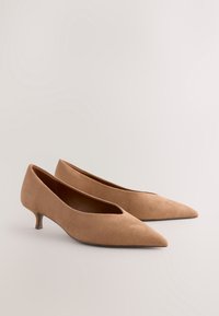 Chaussures à talons hauts en daim beige avec un bout pointu, un petit talon haut et un design latéral ouvert, présentant une texture lisse et des lignes épurées.