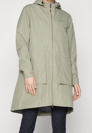 Person iført en beige knælængde vandtæt jakke med hætte, forreste lynlås, to store lommer og sorte bukser.