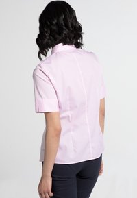 Femme aux cheveux noirs mi-longs portant un chemisier rose clair à manches courtes et un pantalon foncé, debout de dos sur un fond uni.