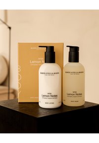 Due bottiglie di gel doccia e lozione, bianche e traslucide, con pompetta nera. Presentano etichette "Note di Limone" e una scatola dorata sullo sfondo.
