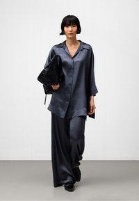 Luftiges, dunkelblaues Satinhemd mit passenden, weiten Hosen. Das Hemd hat einen Kragen und Knöpfe; das Model hält eine schwarze Tasche.