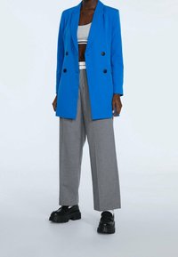 Blazer bleu vif croisé à boutons noirs, pantalon gris à jambes larges, et chaussures plateformes noires épaisses. Design minimaliste, tissu lisse.