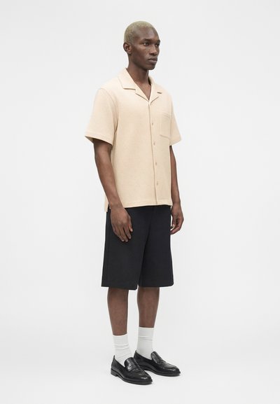 Άνδρας με κοντά ξανθά μαλλιά που φοράει μπεζ κοντομάνικο πουκάμισο με κουμπιά, μαύρα σορτς μέχρι τα γόνατα, άσπρες κάλτσες και μαύρα μοκασίνια, όρθιος.