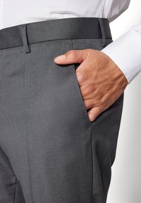 Un pantalon taillé gris avec une texture lisse, doté d'une poche latérale et d'une ceinture élégante, associé à un poignet de chemise blanc.
