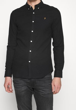 Homme portant une chemise noire ajustée à boutons blancs et un petit logo jaune brodé sur la poitrine, associée à un jean gris foncé.