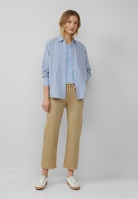 Gestreept blauw overhemd met knopen over een lichtblauw topje, gecombineerd met beige wijde broekspijpen en witte sneakers met een bruine zool.