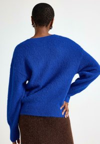 Modrý oversized svetr vyrobený z texturované pleteniny, s kleslými rameny a žebrovaným lemem. Nosený přes hnědou upnutou sukni.