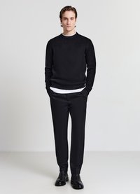 Schwarzer, strukturiert gestrickter Pullover kombiniert mit maßgeschneiderten schwarzen Hosen und schwarzen Lederstiefeln. Unter dem Pullover ist ein weißes Shirt mit Rundhalsausschnitt sichtbar.