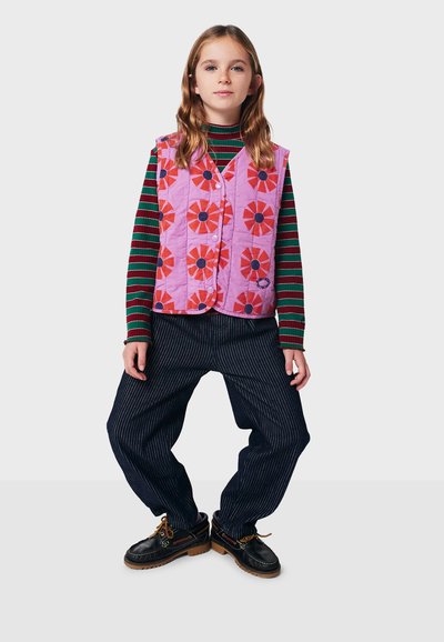 Gestepptes Weste in Helllila mit rotem Blumenmuster, über einem gestreiften Langarmhemd getragen. Weit geschnittene marineblaue Streifenhose und schwarze Schuhe.