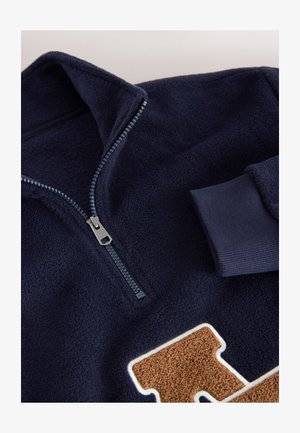 Pulover din fleece bleumarin cu guler cu fermoar pe jumătate, manșete striate și o patch maro texturat în formă de literă pe față. Material moale și cald.