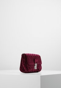 Bolso de mano de terciopelo burdeos acolchado con un cierre plateado, forma rectangular, textura suave y cierre con solapa. Diseño compacto y estructurado.