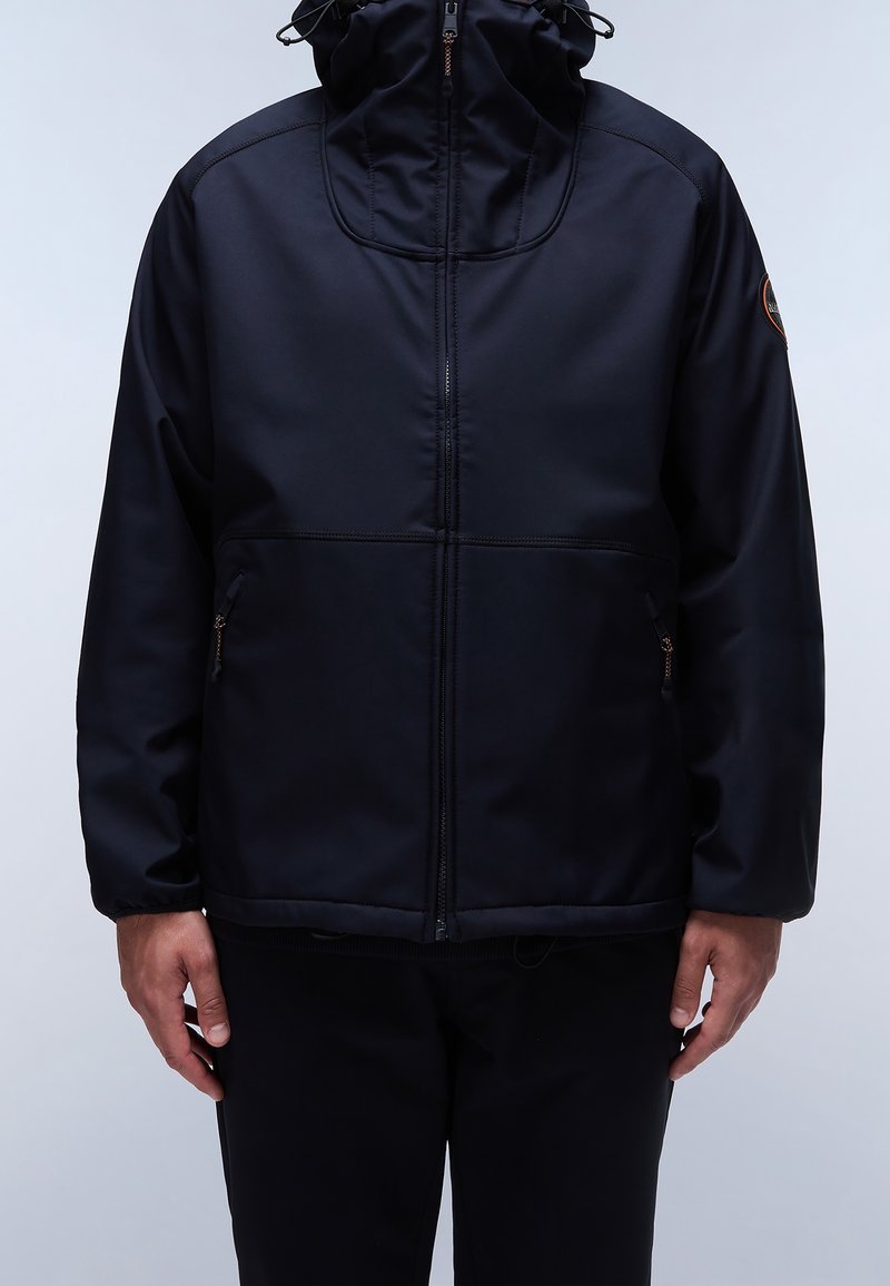 Homme portant une veste à capuche noire zippée avec des poches latérales zippées et un patch logo sur la manche supérieure gauche, debout devant un fond uni.