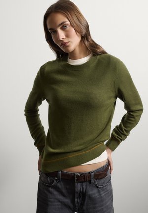 Young woman with long brown hair wears olive green sweater over white shirt and dark jeans with brown belt, standing with hands on hips.