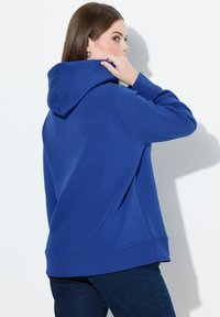 Niebieska bluza z kapturem, dużym kapturem, ściągaczami na rękawach i u dołu, wykonana z miękkiego materiału. Krój luźny, z gładką teksturą.