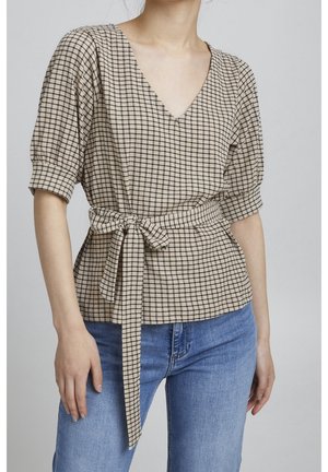 Kvinde iført beige ternet V-halsbluse med bundet bælte i taljen og blå jeans mod ensfarvet baggrund.