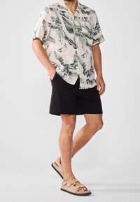 Camisa de manga corta color beige claro con estampados de hojas verdes, combinada con pantalones cortos negros y sandalias marrones con doble hebilla. Atuendo casual.