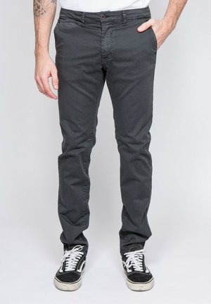 Homme portant un pantalon ajusté gris foncé et des baskets noires et blanches, debout avec une main dans la poche devant un fond uni.