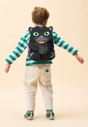Child with light brown hair wears green-and-white striped shirt, beige pants, gray shoes, and black panther-shaped backpack, standing with arms outstretched.
