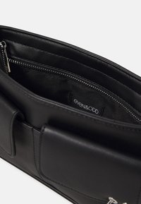 Borsa in pelle nera con texture liscia, tasca interna con zip e etichetta "even&ODD". Presenta un design minimalista e un profilo snello.