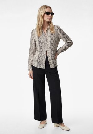 Vrouw met zwarte zonnebril, overhemd met slangenhuidprint en knopen, zwarte wijde broekspijpen en crèmekleurige schoenen met lage hak, staand met één hand op haar heup.