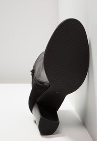 Bottine noire à talon carré, semelle texturée et fermeture éclair latérale, positionnée semelle vers le haut contre un mur blanc et une surface blanche.
