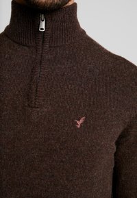 Brun stickad tröja med hög krage, halvdragkedja och en liten rosa logotyp på bröstet. Mjuk textur med ribbat mönster.