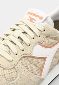 Beige suede sneaker with mesh sections, white branding, and peach accents. Features white laces, a rounded toe, and textured surface.