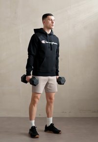 Felpa nera con logo bianco, pantaloncini beige chiaro, in mano manubri esagonali neri. Scarpe da ginnastica e calzini bianchi completano l'outfit. Sfondo neutro.