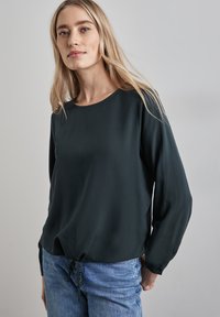 Dunkelgrünes Langarm-Bluse mit rundem Ausschnitt und Kordelzugsaum, aus leichtem Stoff hergestellt. Kombiniert mit blauen Jeans.