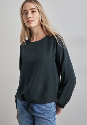 Dunkelgrünes Langarm-Bluse mit rundem Ausschnitt und Kordelzugsaum, aus leichtem Stoff hergestellt. Kombiniert mit blauen Jeans.