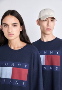 Sudadera de gran tamaño en azul marino con un diseño gráfico que presenta bloques de rojo, blanco y azul, etiquetado como "TOMMY JEANS". Gorro beige claro en el fondo.
