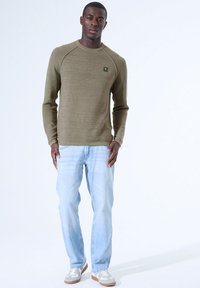 Olivgrünes, strukturiertes Langarmhemd mit Raglanärmeln, kombiniert mit hellblauen Jeans und grauen Sneakers mit weißen Akzenten.