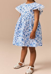 Blau-weißer Blumendruckkleid mit fransigen Ärmeln, gerafftem Taillenbund und vollem Rock, kombiniert mit weißen Riemchensandalen.